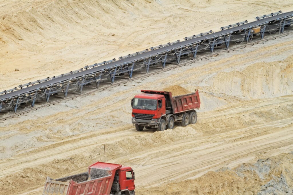 Two red dump trucks transport sand about the expansive quarry. A long conveyor belt runs parallel to the trucks on sandy terrain.