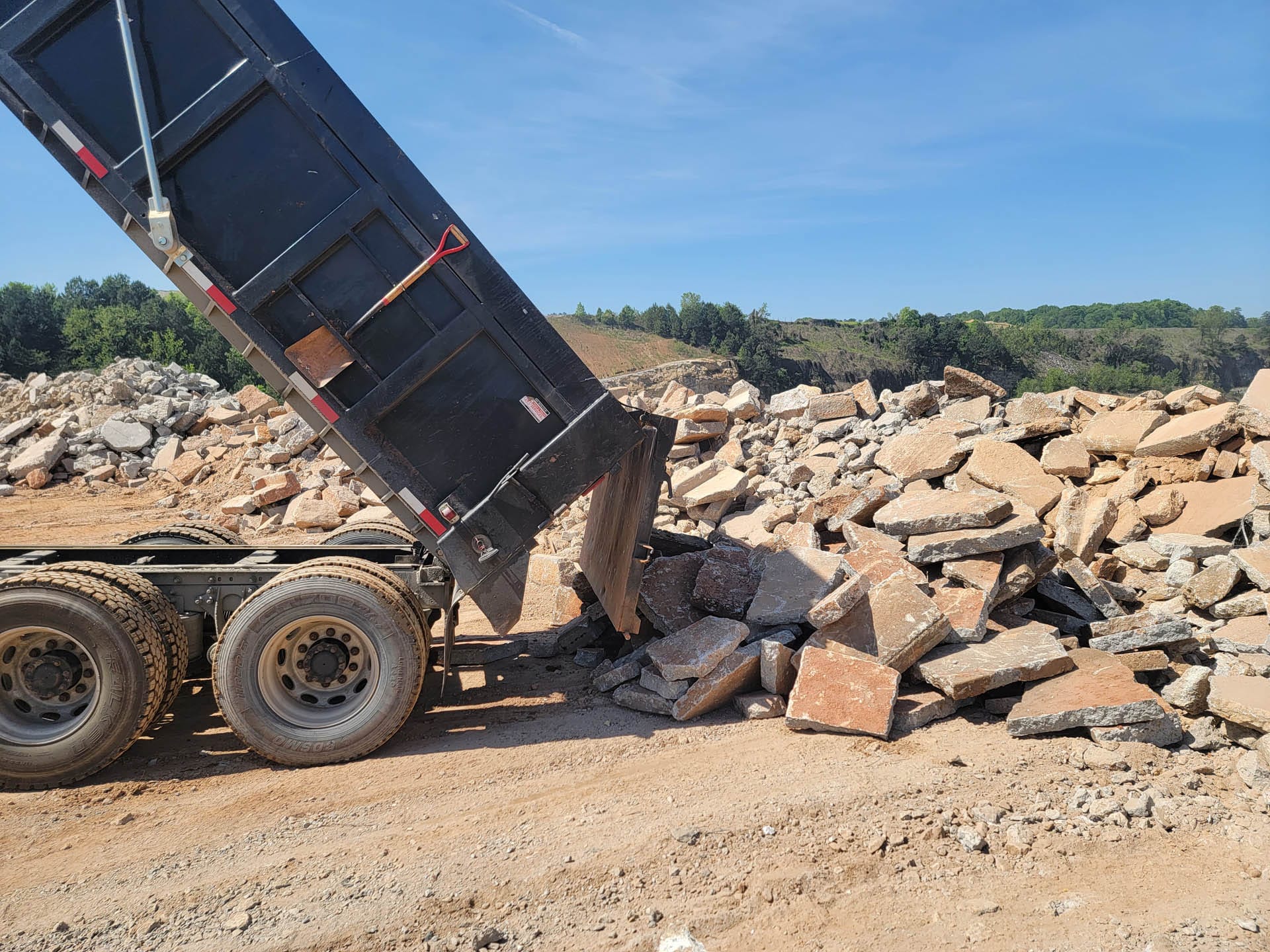 A dump truck unloading rubble onto a pile at a construction site with trees in the background.