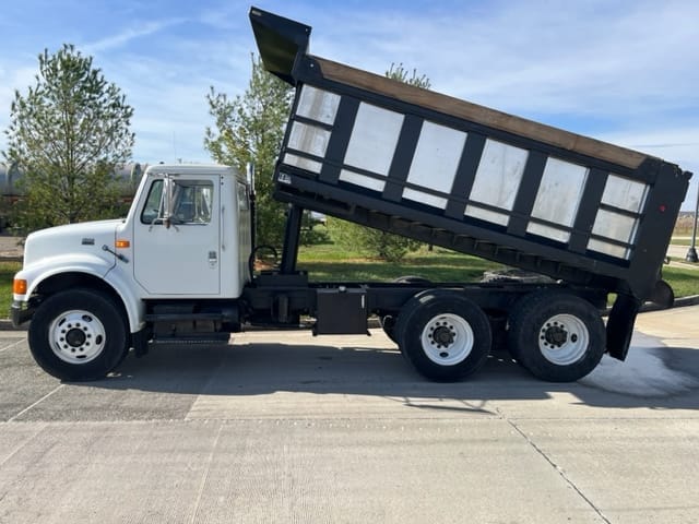 About a white dump truck with its bed raised on a sunny day, it stands proudly parked on a concrete surface with trees providing a lush backdrop.