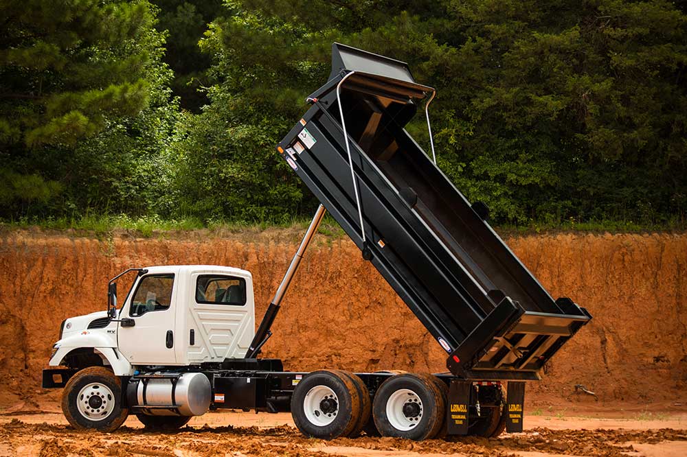 A white dump truck with its bed raised stands ready on a dirt ground, framed by trees in the background, symbolizing reliable services.